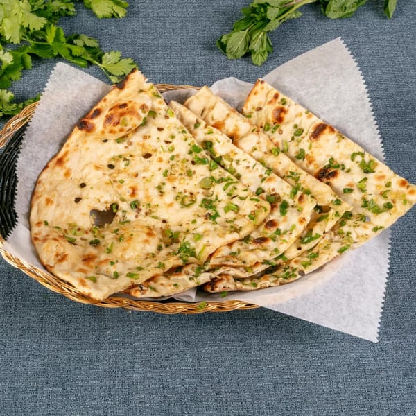 Golden-brown naan bread, garnished with green herbs, served in a woven basket on a blue tablecloth.