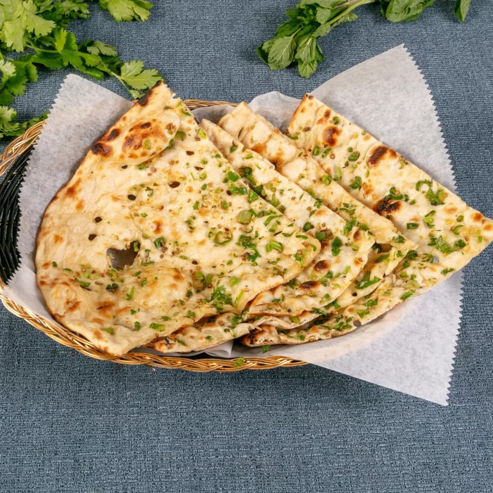 Golden-brown naan bread, garnished with green herbs, served in a woven basket on a blue tablecloth.