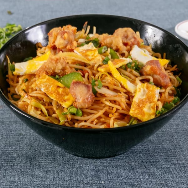 Noodles with fried chicken, scrambled egg, and green onions in a black bowl.