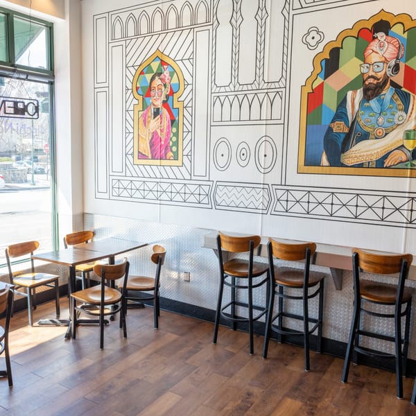 Restaurant interior with colorful Indian-inspired murals, wooden tables, and bar seating along a window.