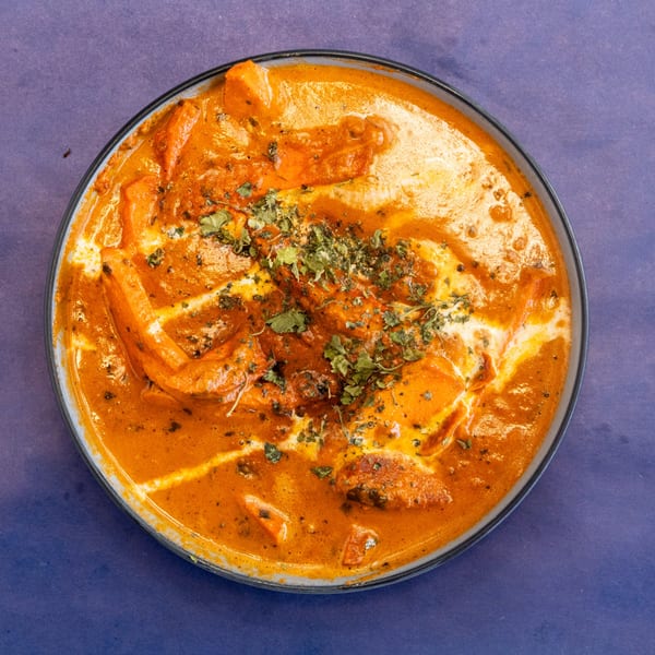 Overhead view of a bowl of orange butter chicken with cream swirls and green herbs.