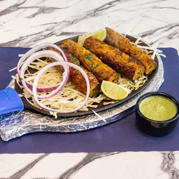 Four seekh kebabs with sliced onions, lime wedges, and green chutney on a hot plate.