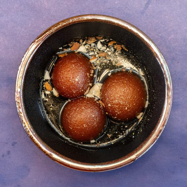 Three gulab jamun in syrup with chopped nuts, in a dark bowl on a purple surface.