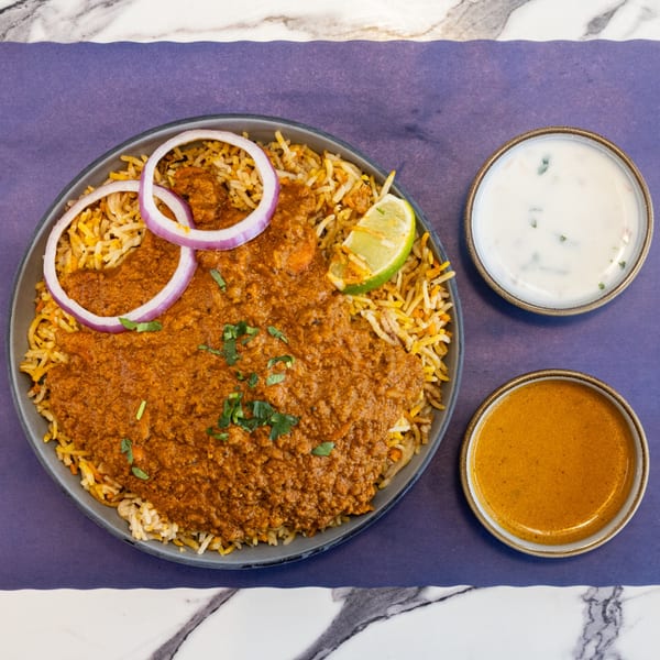 Chicken biryani with red onion, lime, and two sauces on a purple and marble background.