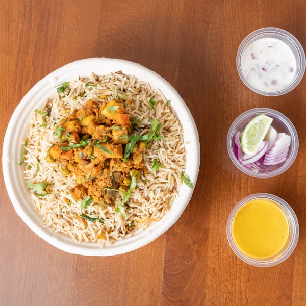 Vegetable biryani in a white bowl with sides of raita, sliced red onions, lime, and curry sauce.