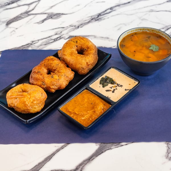 Three golden-brown medu vada on a black plate with two chutneys and a bowl of sambar on a blue mat.