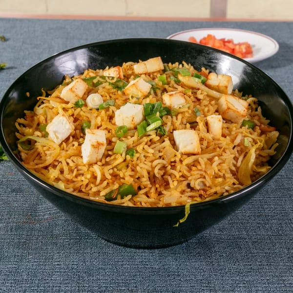 A black bowl filled with paneer fried rice, garnished with green onions, on a blue textured surface.