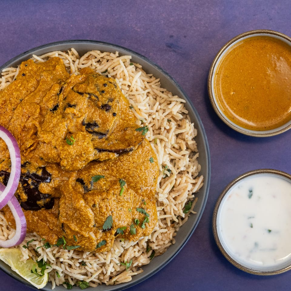 Eggplant curry with rice, red onion, lime, and two side sauces on a purple background.