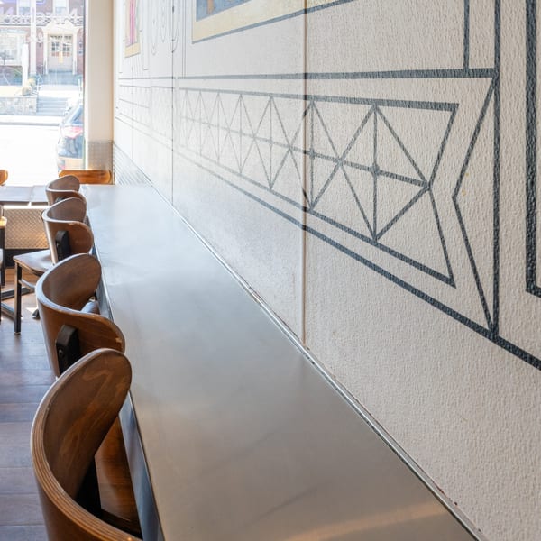 Long bar-height table with several wooden chairs and a patterned wall in a bright restaurant.