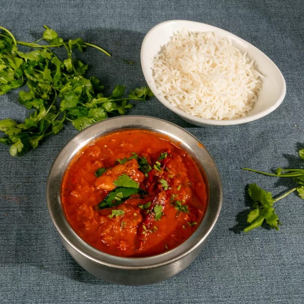 A metal bowl of red curry with cilantro and a white bowl of white rice on a blue surface.