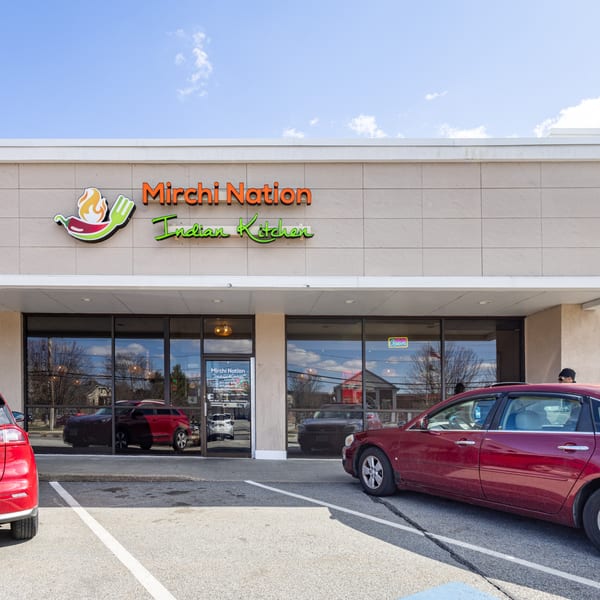 Mirchi Nation Indian Kitchen storefront with its logo, large windows, and parked cars in front.