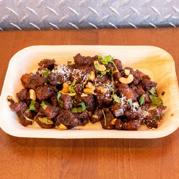Dark brown chicken dish with cashews, cilantro, and shredded coconut on a light wooden plate.