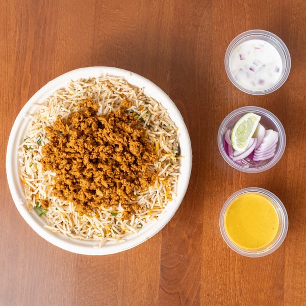 Keema biryani with rice, ground meat, and sides of yogurt, onions, and sauce on a wooden table.