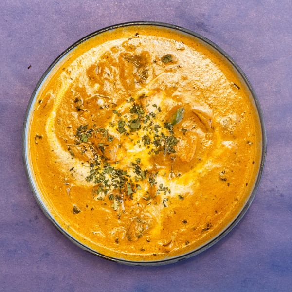 Overhead view of a bowl of orange curry with white cream swirls and green herbs.