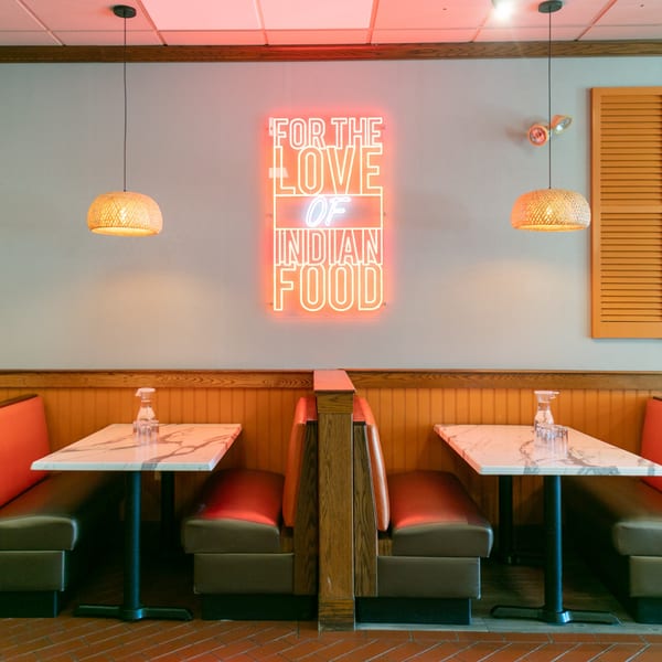 Restaurant interior with a neon sign reading "FOR THE LOVE OF INDIAN FOOD" above two booths.